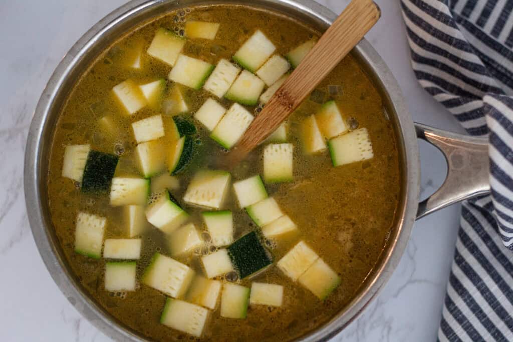 Chopped zucchini simmering in broth with a wooden spoon.