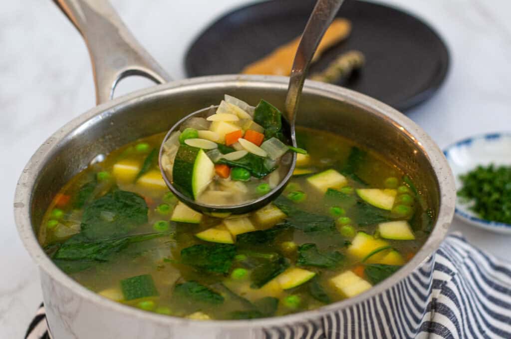 A ladle lifting soup, showing orzo and vegetables.