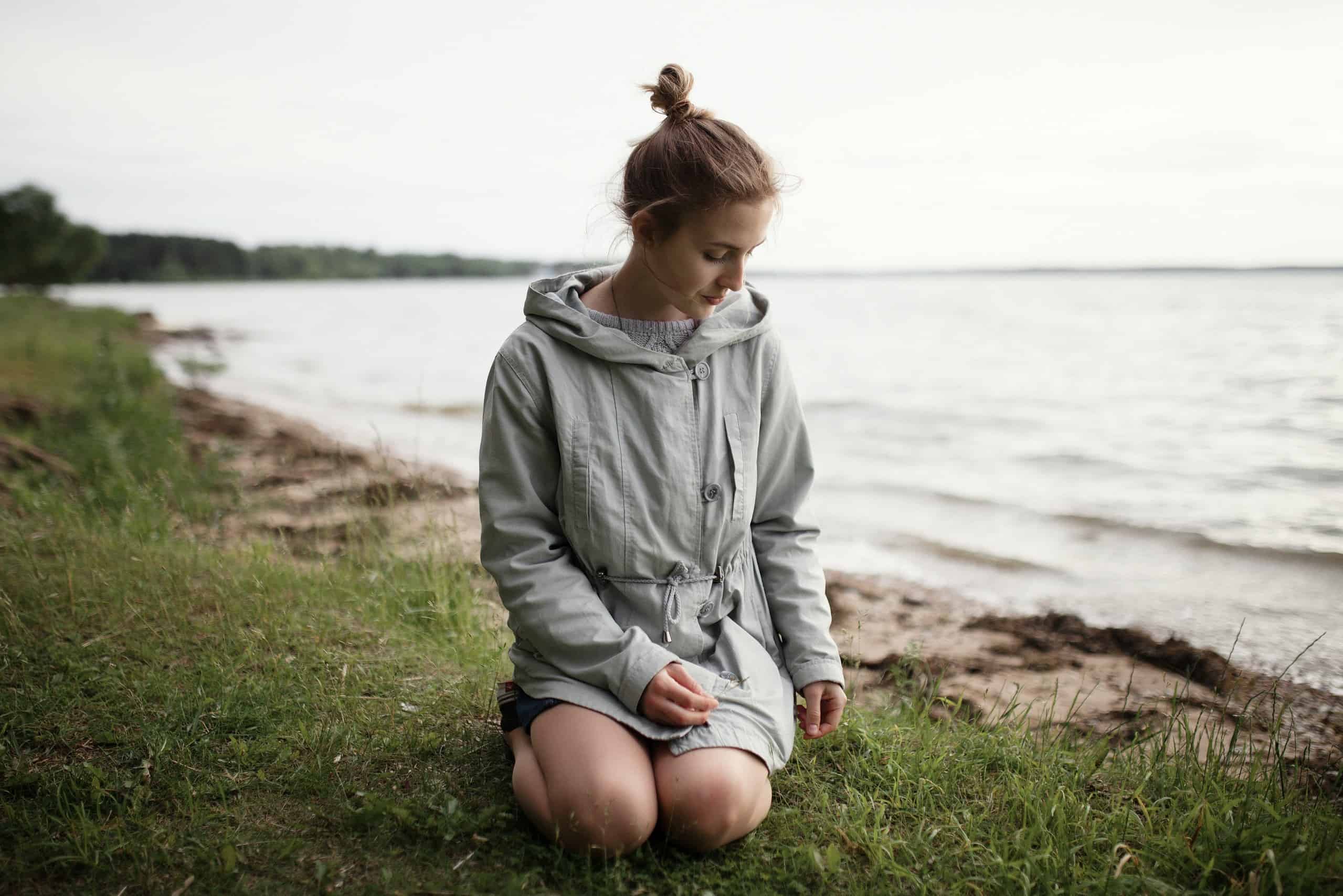Peaceful woman praying outdoors near a lake or river, kneeling on grass with eyes closed in reflecti.