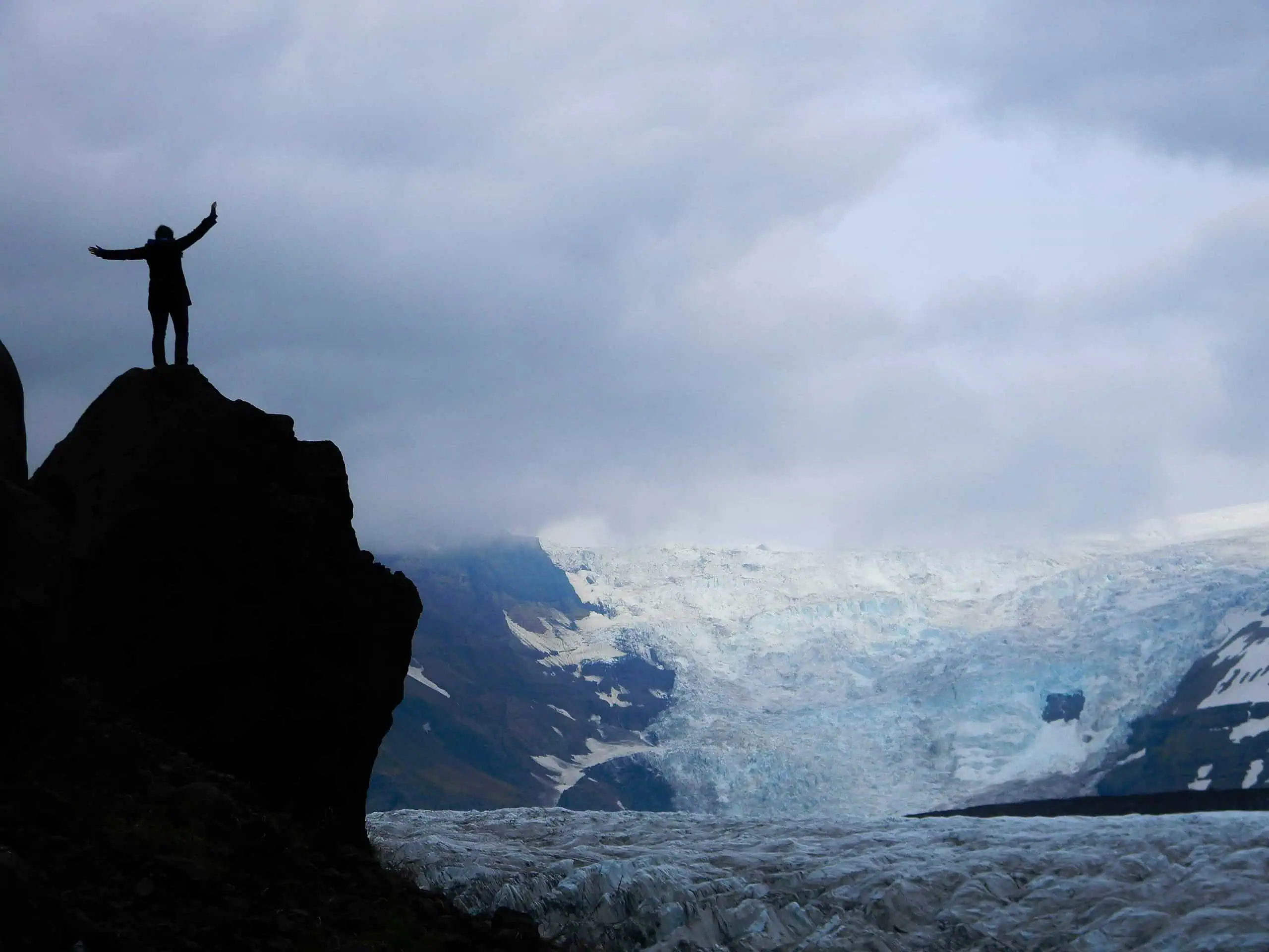 Person standing triumphantly on a mountain peak with arms raised, overlooking a vast glacier and rug.
