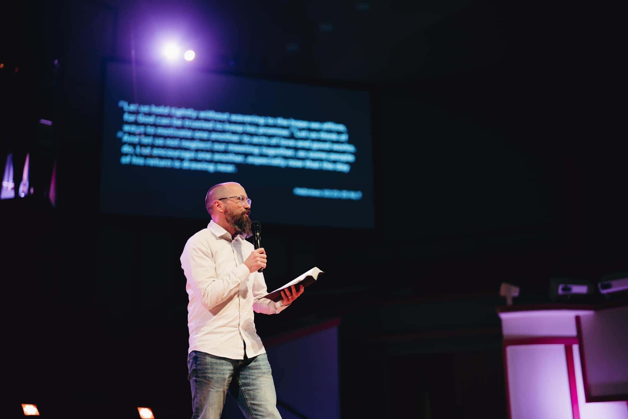 Worship leader speaking at NLW International event with a microphone and screen in the background.