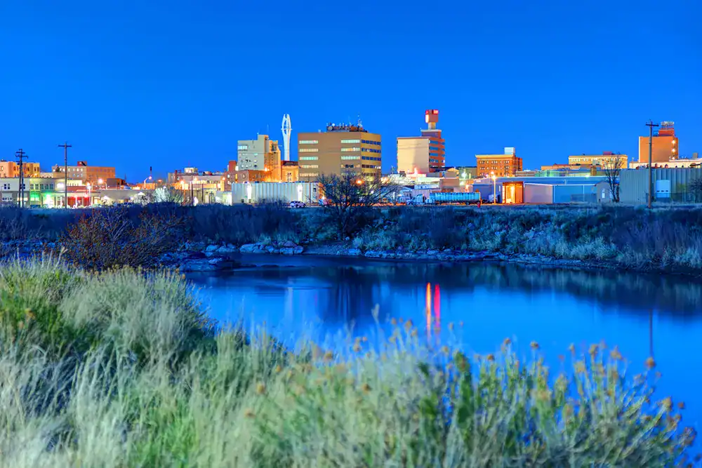 City skyline with illuminated buildings and calm river in the foreground.