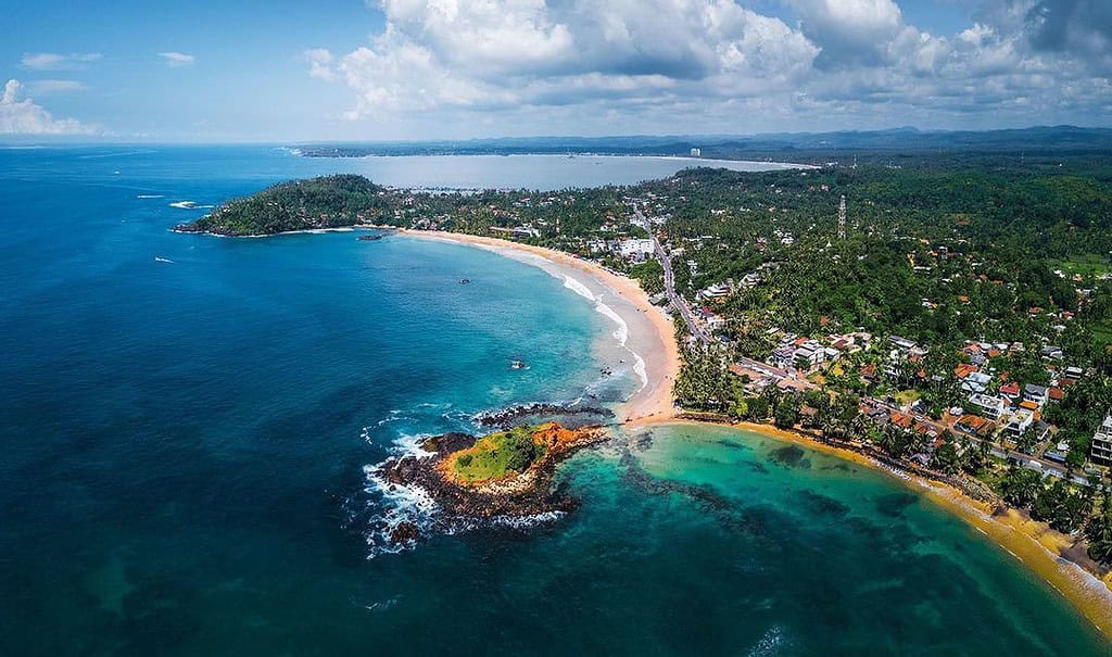 Aerial view of tropical coastline with beach, ocean, and green hills, showcasing natural beauty and vibrant community.