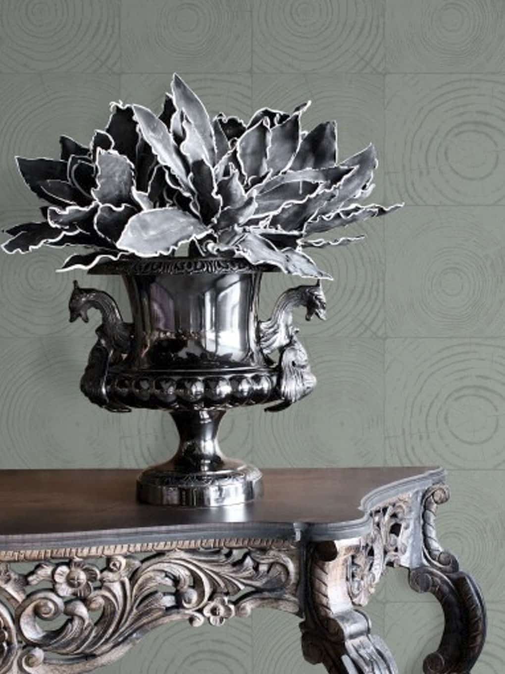 Silver decorative vase with lush green plant on ornate wooden table.
