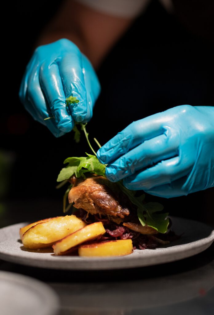 Chef's hands in blue nitrile gloves plating a gourmet dish.