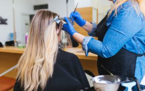 Hair stylist wearing blue disposable gloves applies hair color to client's blonde hair.