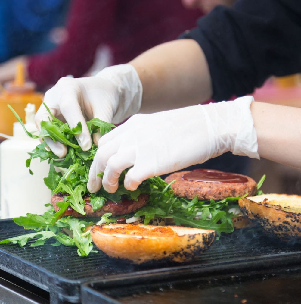 Close-up of gloved hands adding arugula to gourmet burgers on a grill.