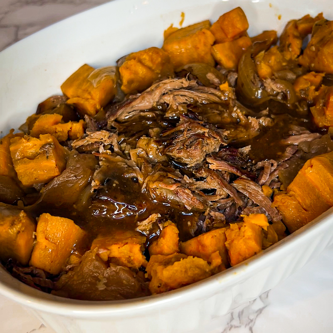 Easy Pot Roast with Sweet Potatoes: A Nourishing, Faith-Filled Family Favorite | The Not so Modern Housewife A hearty pot roast with sweet potatoes, featuring tender shredded beef surrounded by large chunks of orange sweet potatoes and onions in a rich, savory gravy, served in a white casserole dish on a marble countertop.