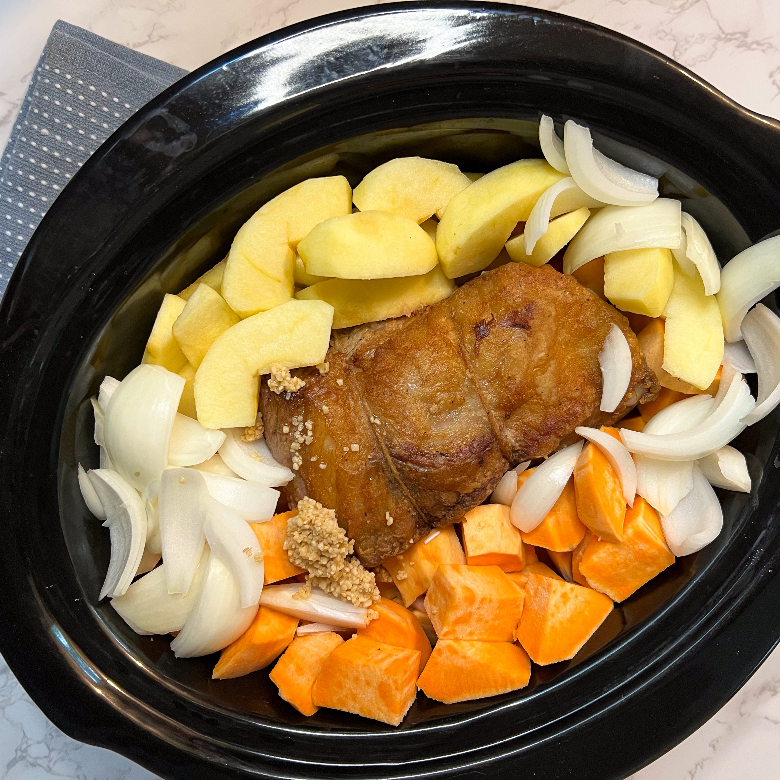 Easy Pot Roast with Sweet Potatoes: A Nourishing, Faith-Filled Family Favorite | The Not so Modern Housewife Uncooked pot roast with sweet potatoes in a black slow cooker, featuring a seared beef roast surrounded by chopped sweet potatoes, sliced onions, minced garlic, and green apple wedges, ready for a slow and savory cook.