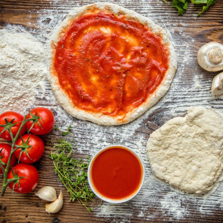 The Ultimate Guide to Mastering Sourdough Pizza Crust: Tips & Tricks | The Not so Modern Housewife pizza dough rolled out and covered in sauce on a wood surface dusted with flour - surrounded by pizza ingredients