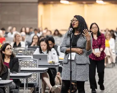 woman standing at a mic with rows of tables next to and behind her with people sitting at them