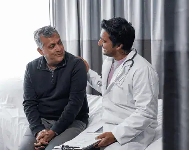 two men sitting and talking, one appears to be a doctor, in a lab coat. the doctor has his hand on the man's shoulder