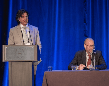Man standing at a podium and another man is next to him at a table, talking into a mic