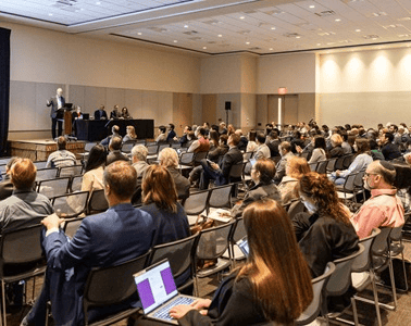 room with rows of chairs, filled with people. there is a speaker at the front on a stage