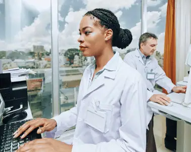 woman sitting at a desk, hands on keyboard. there is a man behind her, also at a desk. there is are large windows behind them