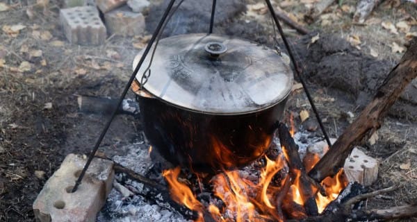 Showing a metal camp oven suspended over an open fire, cooking lamb outdoors.
