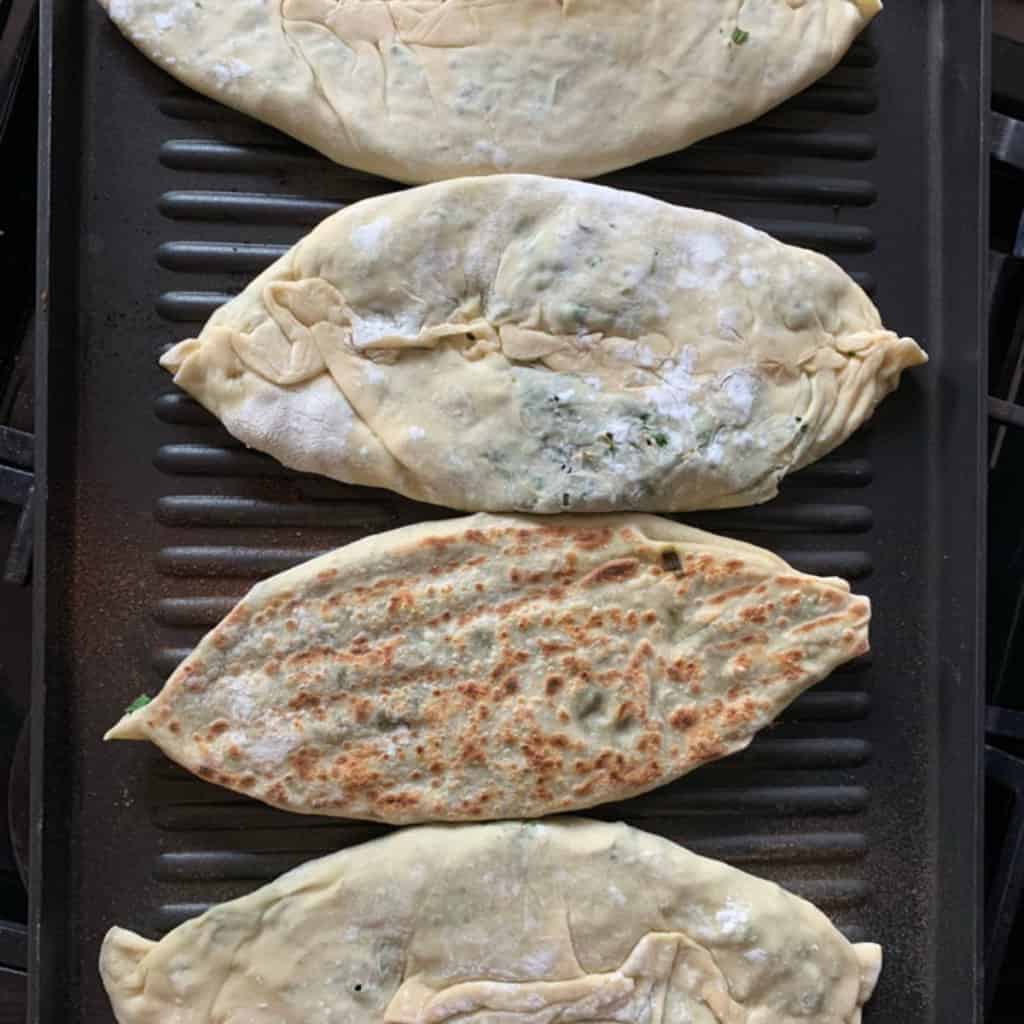 Two Jingalov Hats cooking on a hot cast-iron griddle, seam side down, developing charred blisters on the surface.