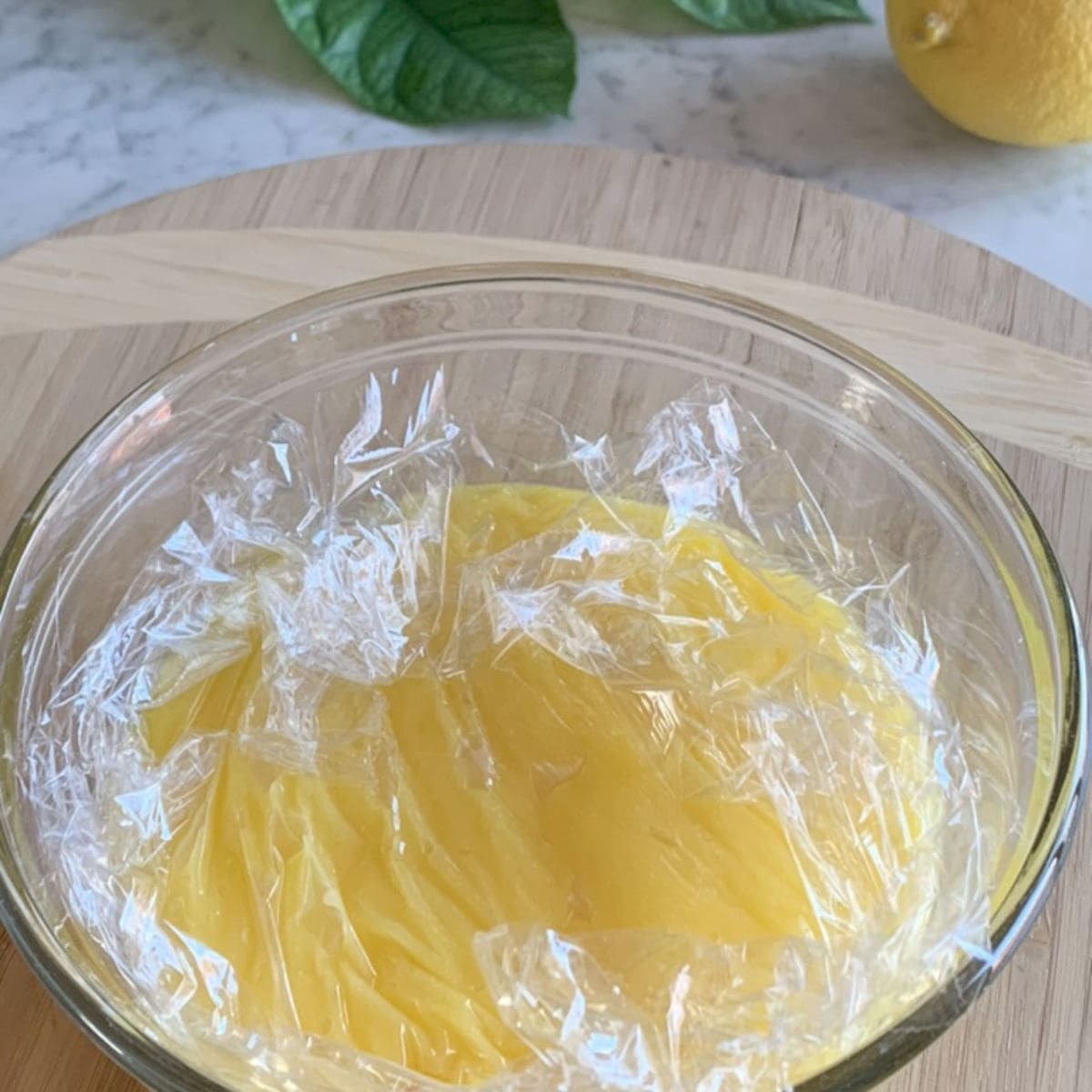 Lemon curd in a glass bowl with the plastic film over it.