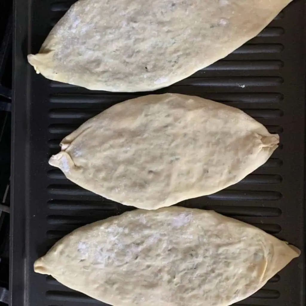 Three Jingalov Hats cooking on a hot cast-iron griddle, seam side down, developing charred blisters on the surface.