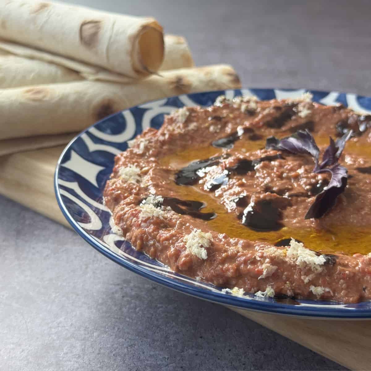 Muhammara in a shallow dish, drizzled with olive oil and pomegranate molasses, with rolled lavash on a board.