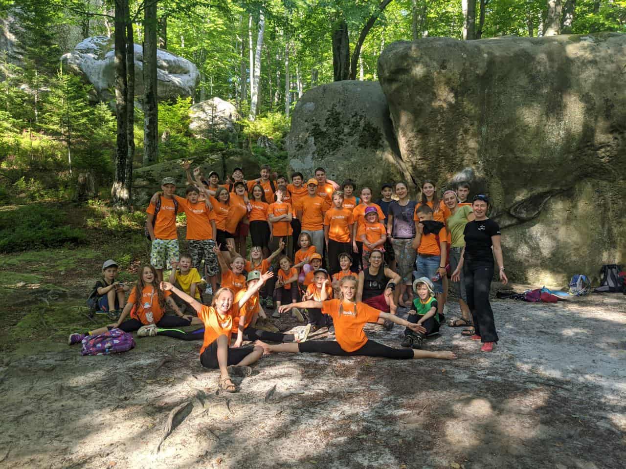 Group of children and adults in outdoor forest setting with rocks and trees.