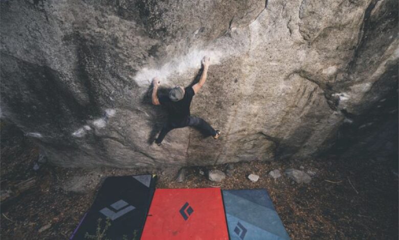 Climber scaling a steep outdoor rock face with crash pads below.