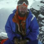 High-altitude mountaineer in blue gear on snowy mountain summit with glacier backdrop.