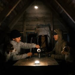Gentleman and woman in outdoor gear sharing flashlight inside rustic wooden cabin.