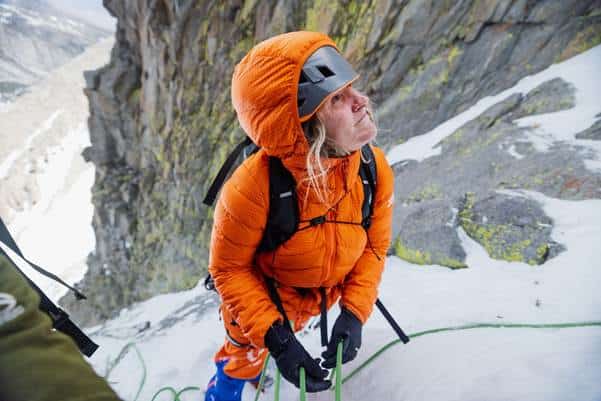 Climber in orange insulated outdoor gear near snow-capped mountain cliffs, engaging in mountain climbing.