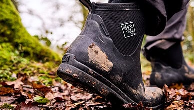 Close-up of muddy Muck Boots on a forest trail during outdoor activity.