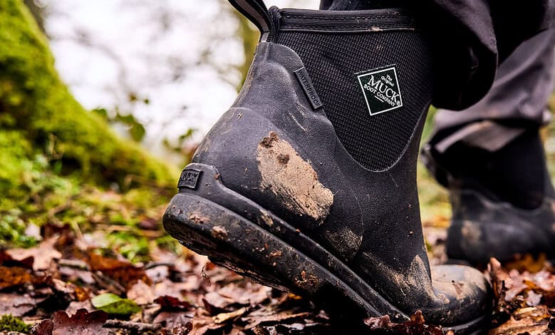 Close-up of muddy Muck Boots on a forest trail during outdoor activity.