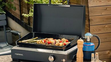 Portable outdoor grill with food and accessories on a garden table.
