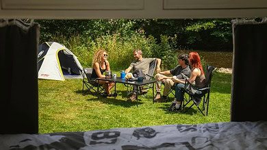 Friends enjoying outdoor cooking and dining by a river with Campingaz grill and tent.