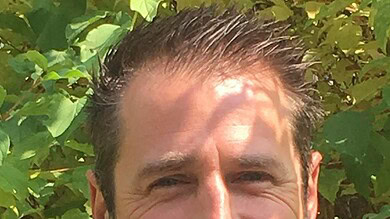 Smiling man outdoors with greenery background, professional headshot.