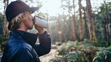 Person enjoying outdoor drink in forest during daytime, wearing outdoor gear.