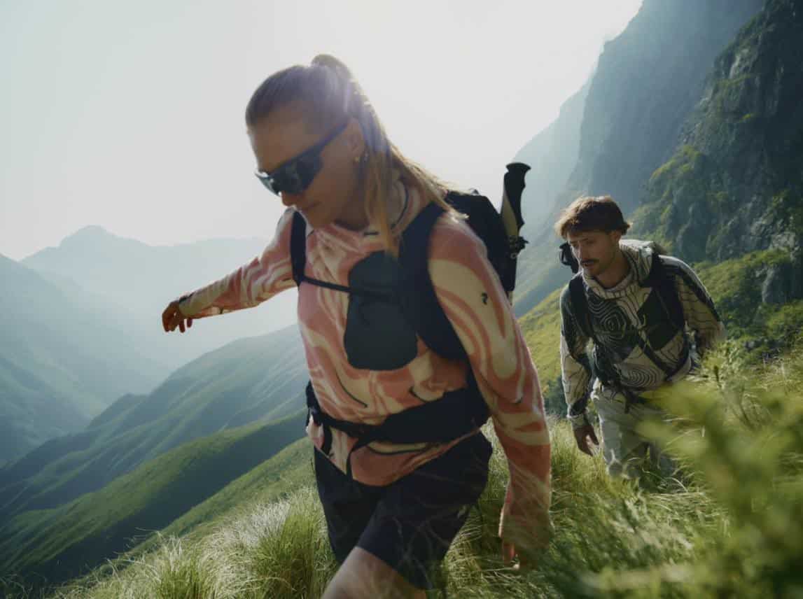 Two hikers trekking through lush green mountains with backpacks and outdoor gear.