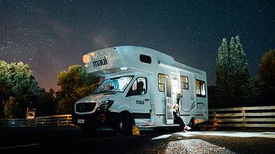 Motorhome parked under a starry night sky with trees and a wooden fence nearby.