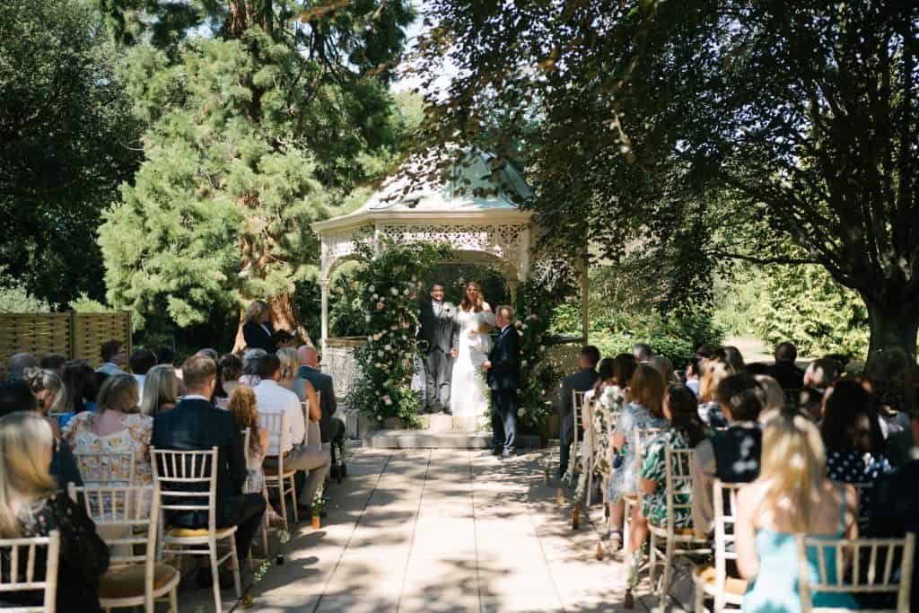 Wedding ceremony outdoors at Quex Park with guests seated under trees.