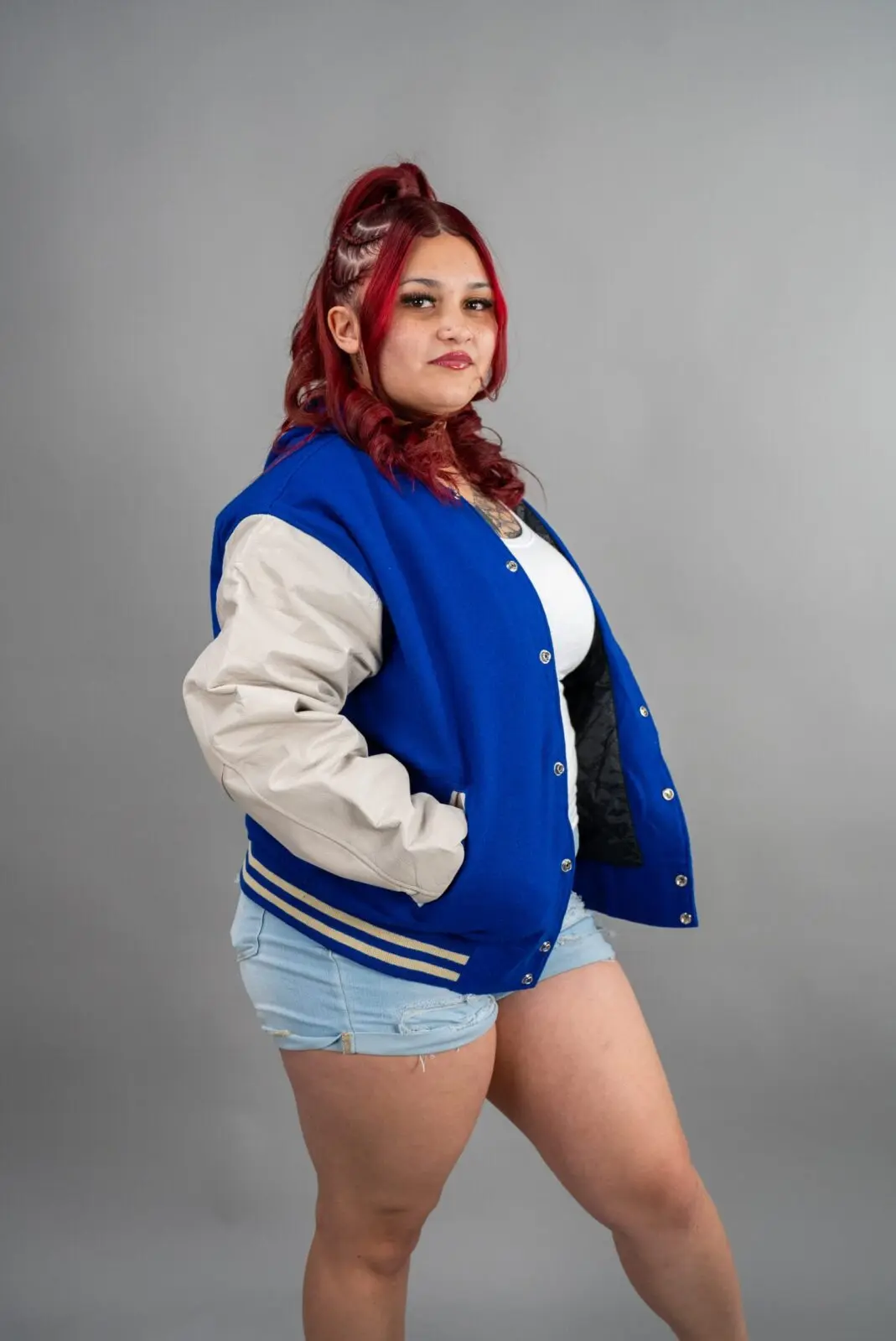 Woman with red hair wearing a blue varsity jacket and denim shorts against gray background.