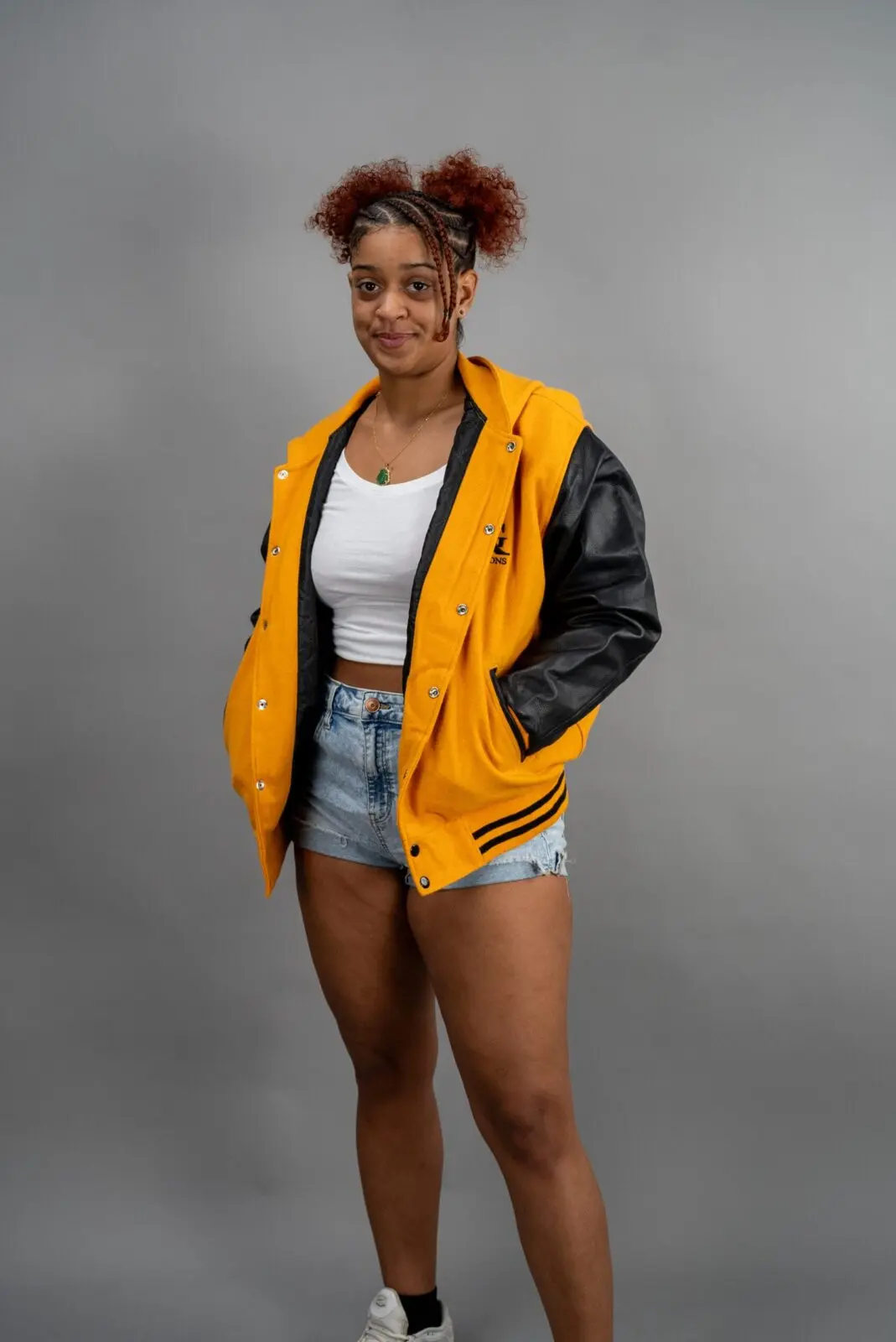 A woman wearing a yellow and black Reign Gear LLC jacket, white crop top, and denim shorts standing.