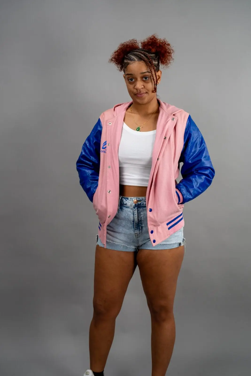 Young woman in casual pink and blue varsity jacket with denim shorts against gray background.