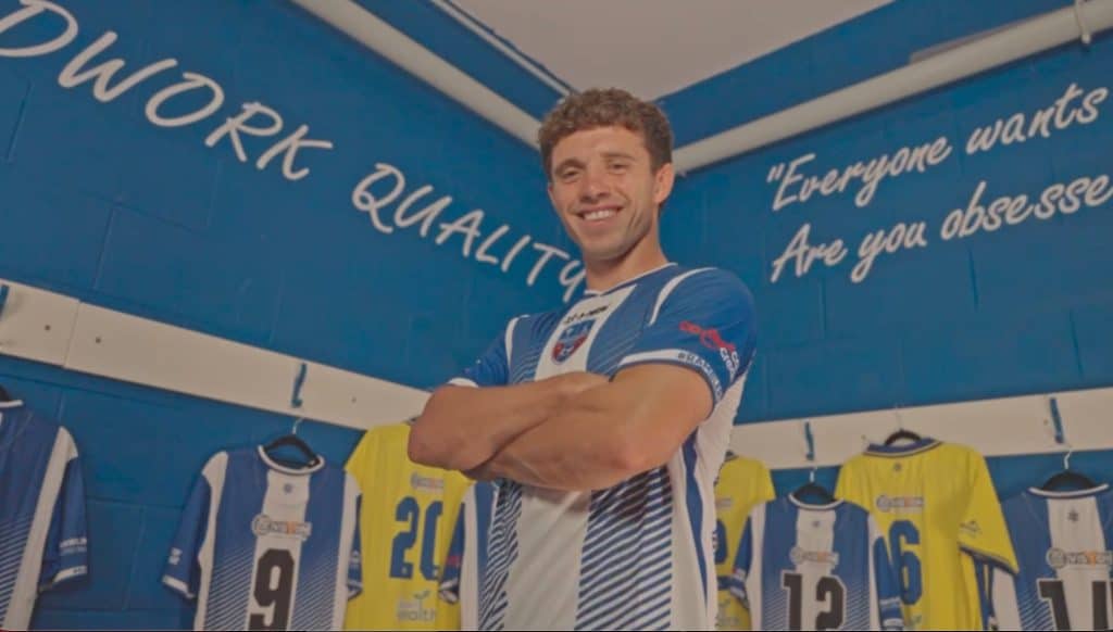 Sandbach United player smiling in the kit room with team jerseys behind him.