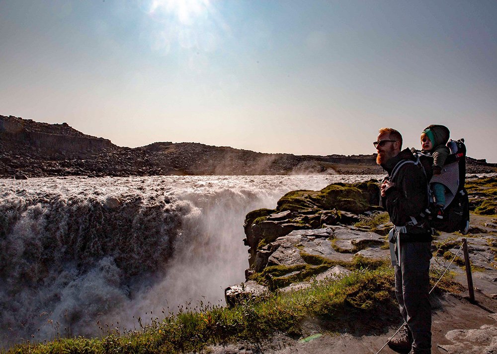 A father carrying his child in a back carrier while standing next to the powerful Selfoss waterfall, with mist and sunlight in the background.