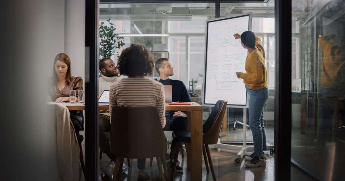 A diverse group of people gathered indoors, looking attentively at a whiteboard. The scene includes both men and women wearing casual clothing, with natural light coming in from a nearby window.