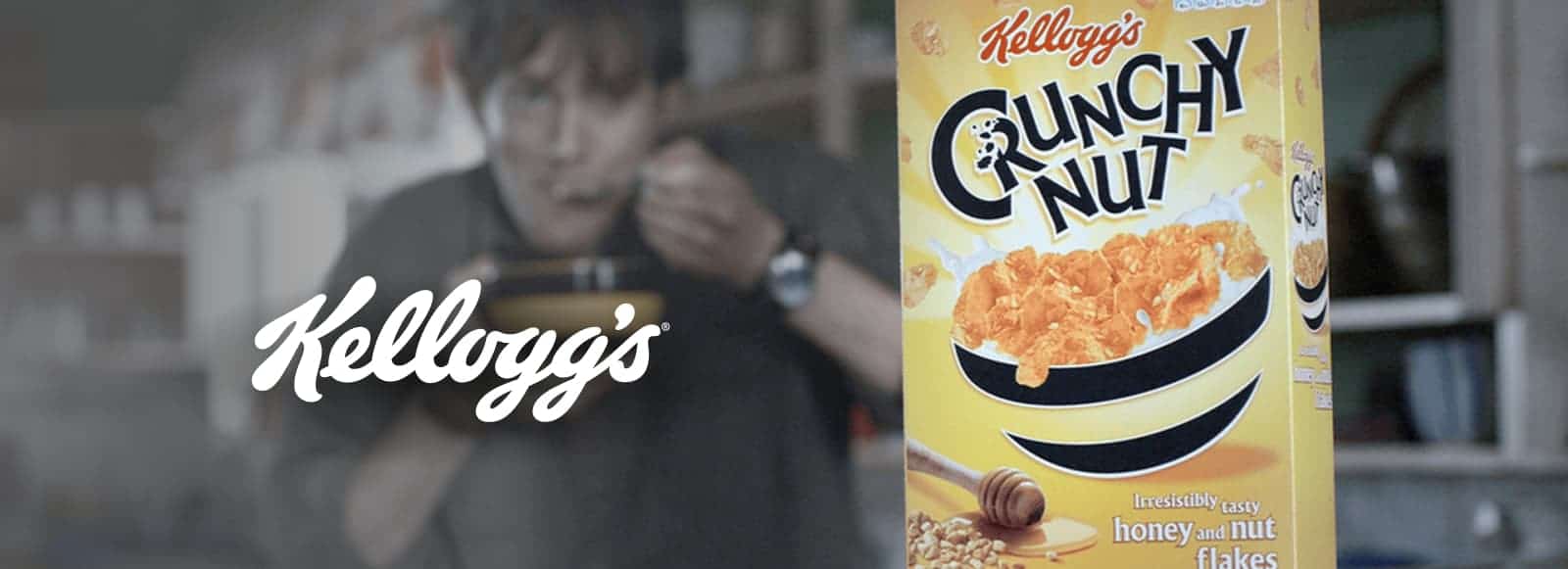 A person sitting indoors, enjoying a bowl of cereal made from honey nut flakes, with a box of Kellogg's CRUNCHY NUT nearby.