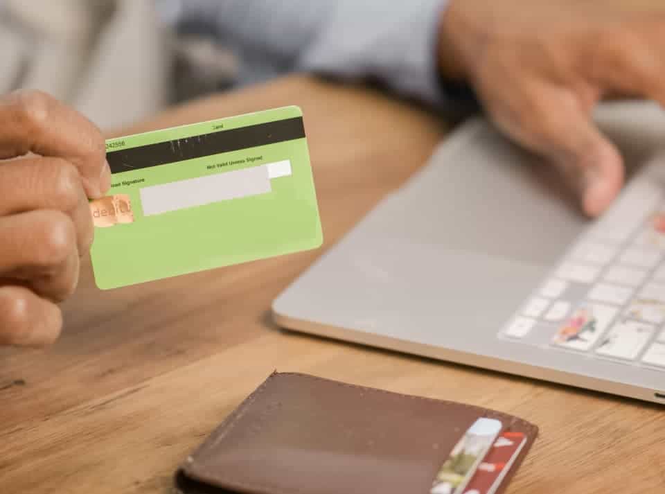 A person holding a credit card in an indoor setting with office supplies, a laptop, and a tablet computer on the table.