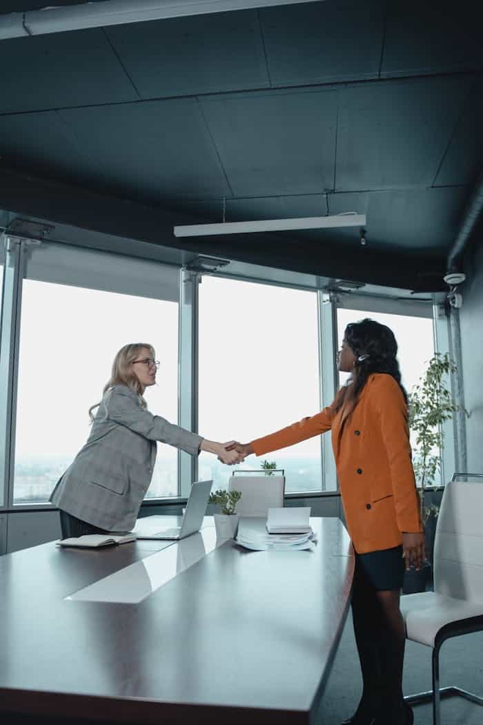 Two businesswomen shake hands across a conference table in a modern office setting.