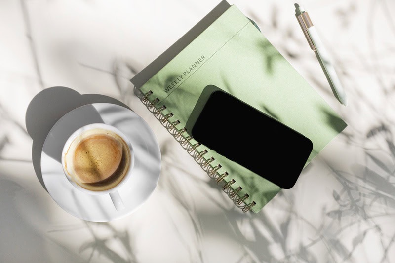An green journal on a white desk next to a cup of coffee with a phone on top. Ready to learn about Self-care Gal
