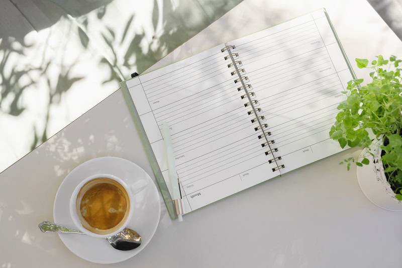 An open journal on a white desk with a coffee and a plant next to it. Someone is taking notes while they learn about Self-care Gal
