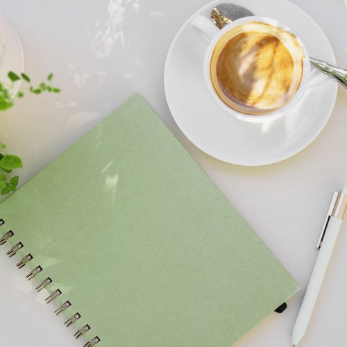 A green spiral notebook, a white pen, and a cup of coffee on a saucer are arranged on a white surface, with some green plant leaves visible in the corner—perfect for moments of productive self-care.